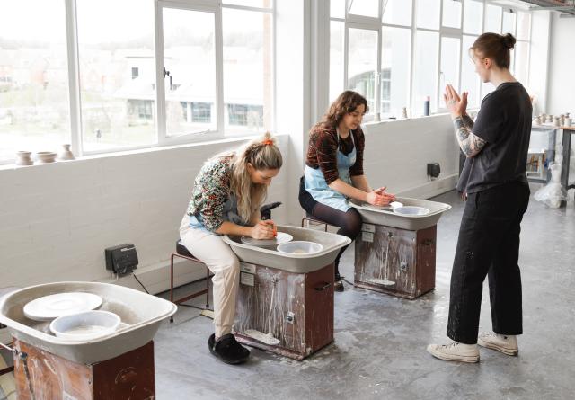 image of clay studios at world of wedgwood, with two women sat at a potter's wheel each and an instructor stood infront. 